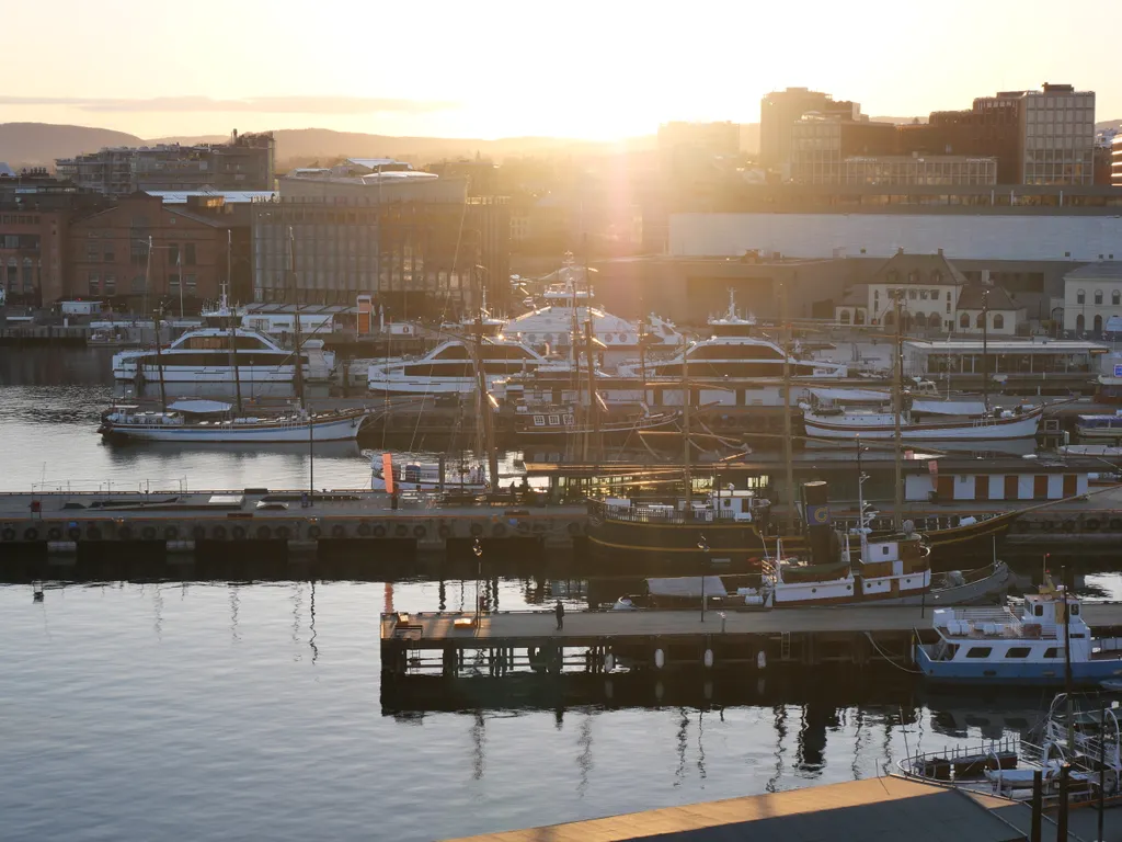 Hinter dem Hafen von Oslo sieht man die Sonne Untergehen. Am Hafen sind einige Schiffe an den Stegen zu sehen.